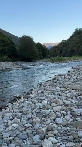 A clear, wide shot shows a fast-flowing river with a rocky shore. The water is a bright turquoise color and appears to be moving swiftly. The riverbanks are lined with lush green trees and foliage, and in the distance, mountains rise against a clear blue sky. The overall scene is serene and picturesque, with a focus on the natural beauty of the landscape.