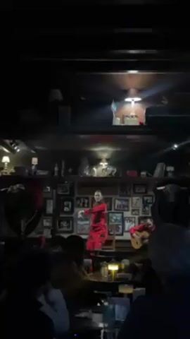 The video shows a dimly lit, crowded bar with a small stage where a flamenco dancer and guitarist are performing for an audience. The dancer, wearing a red dress, stands front and center, while the guitarist sits to the side. The background is cluttered with pictures, bullfighting memorabilia, and other decor. The audience is seated at tables, watching the performance.