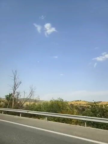 The video shows a roadside view from a moving vehicle on a sunny day. A clear blue sky with a few scattered clouds is visible. In the foreground, a highway barrier runs along the side of the road, and the asphalt road itself is visible. Beyond the barrier, there is a dry, grassy landscape with sparse vegetation, including some green bushes and a few dead, bare trees. The camera appears to be panning slightly or the vehicle is moving, causing a slight motion blur. No people or specific objects are clearly discernible other than the landscape features and the road infrastructure.