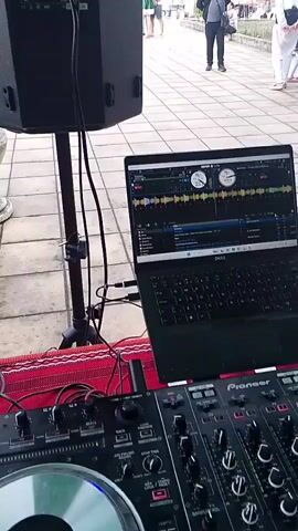 The video captures an outdoor cultural event in Mogila, Bulgaria, featuring traditional folk dancing. The scene opens with a close-up of a DJ setup, including a black Dell laptop displaying DJ software, a Pioneer mixer, and a large black speaker on a stand. A man walks in the background.

The camera then pans across a paved outdoor area, revealing several people, including children, gathered and dancing. Many participants are dressed in vibrant traditional Bulgarian folk costumes, primarily red dresses with white aprons, black vests, and white shirts. Some are dancing in a line or circle, characteristic of a "hora" folk dance.

In the foreground, there is a large, grey decorative urn with a plant. A black metal staircase with a railing is also visible. The setting includes trees and what appear to be public buildings in the background. A green awning provides shade over part of the area. The event appears to be taking place during the daytime, suggesting clear weather. The overall atmosphere is festive and lively, with music playing from the DJ setup.