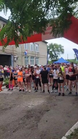 A group of runners are gathered at a starting line for a race. They are standing under a red and white inflatable arch. Many of the runners are wearing athletic clothing and bib numbers. Some are talking to each other, while others are looking ahead. The ground is made of dirt and gravel. In the background, there is a brick building with windows. There are also trees and bushes. One person is holding a phone up to take a picture.