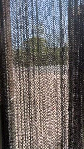 The video captures a close-up perspective through a dark, metallic mesh screen or curtain. Initially, the mesh fills the frame, with a blurred background showing green trees, a light-colored ground surface, and a clear blue sky. A person's hand, with fingers extended, then enters the frame from the right, pushing or reaching through the mesh. The mesh material appears flexible, distorting slightly as the hand interacts with it. Bright natural light suggests it is daytime. The background remains out of focus, emphasizing the texture of the mesh and the hand. No text is visible. The scene depicts an outdoor setting.