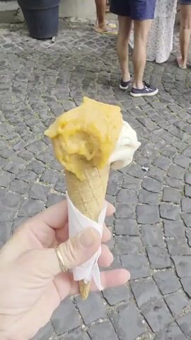 The video shows a person holding a cone with two scoops of gelato, one yellow and one white. The background is a cobblestone street with several people walking around. The scene appears to be in front of a gelateria with a sign that reads "GELATERIA PICCHIO". Some people are queuing to enter the gelateria, while others are walking past. The lighting suggests it is daytime, possibly late morning or afternoon, with bright sunlight. The atmosphere seems relaxed and casual, with people enjoying themselves in what appears to be a tourist area. The overall impression is a pleasant, everyday scene of enjoying gelato on a sunny day in Italy.