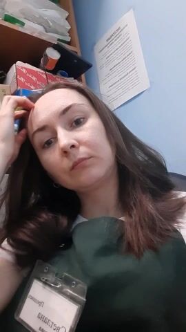 A woman with shoulder-length brown hair and fair skin is centered in a selfie-style video. She has a serious expression. Her hair is partially pulled back behind her ear, and she adjusts it slightly with her hand. She is wearing a white t-shirt covered by a dark green apron, with an ID badge attached to it. The badge is obscured. Behind her is a light blue wall, a wooden bookshelf with various items, including boxes, papers, and a container. A printed document is taped to the wall. The camera angles slightly up towards the woman.