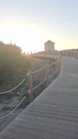 A wooden boardwalk curves to the right, leading towards a small stone windmill silhouetted against a bright, hazy sunset. The sun is low in the sky, casting a warm glow and lens flares across the scene. The boardwalk is bordered by a simple wooden fence with rope railings, separating it from dry grass and low bushes. The sky is clear and pale.