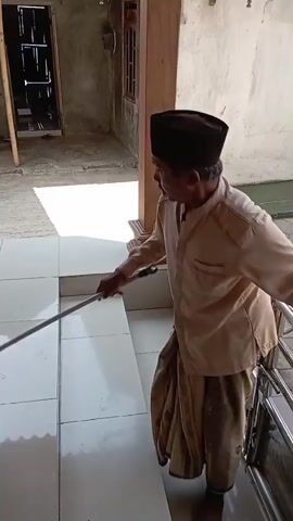 A man wearing a tan shirt, brown patterned sarong, and black cap is sweeping the tiled floor with a broom. He is standing on a raised tiled area with steps leading down. To his left, there is a metal drain grate and a coiled green hose. In the background, there is a motorcycle, clothes hanging on a line, and a building with a corrugated metal roof. The man is actively sweeping, moving the broom across the floor.