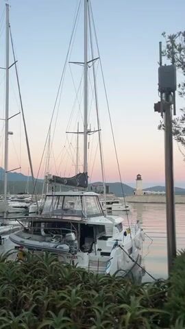 The video pans from left to right across a harbor at sunset. The foreground is dominated by lush green foliage. In the midground, several sailboats and a catamaran are docked. The catamaran in the center is white with black sails and has a tender attached to its side. A person in a yellow shirt is visible on the deck of the catamaran. In the background, a lighthouse stands on a pier. The sky is a gradient of soft pink and blue hues, with the sun just below the horizon. The water is calm and reflects the colors of the sky.