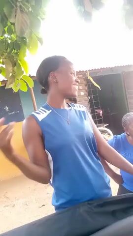 A young, dark-skinned woman, wearing a blue sleeveless top with white stripes on the shoulders and a silver cross necklace, waves at the camera and smiles. She stands outdoors, possibly on a dirt road or yard, with trees overhead providing partial shade. Another woman, also in a blue top, is partially visible behind her. In the background, there's a building with a corrugated metal roof and painted walls, including a sign with the word "Startimes" visible. The camera appears to be handheld, giving a somewhat shaky perspective of the scene.