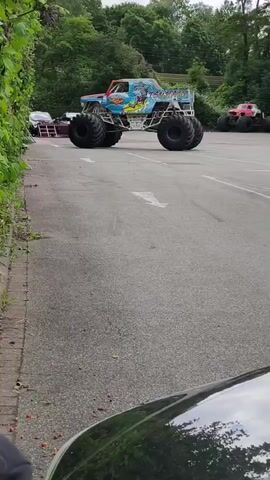 The video shows a monster truck with the name "Sea Serpent" written on its side in blue lettering. The truck is light blue with images of sea creatures on it. It has large, black tires and appears to be parked on an asphalt surface. In the background, there is a red monster truck parked behind the blue one. The area is surrounded by lush green trees and bushes, suggesting an outdoor setting, possibly a parking lot or event space. A portion of a dark-colored vehicle, likely a car, is visible in the foreground at the bottom of the frame. The sky is overcast.