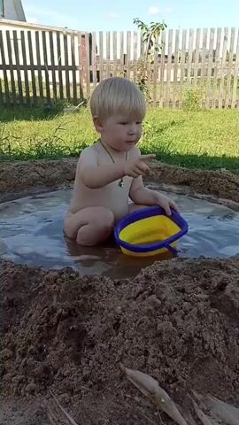 A young boy is playing in a muddy puddle in a backyard. He is sitting in the water with a yellow and blue bucket. The boy has blonde hair and is wearing a green necklace. He is looking down at the bucket with a focused expression. The backyard has a wooden fence in the background and green grass. The sky is blue with some clouds, suggesting it is a sunny day. The overall scene depicts a child enjoying outdoor play.