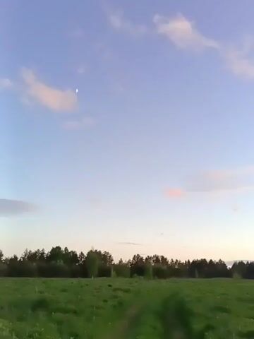 The video shows a green field with a line of trees in the background. The sky is blue with some light pink clouds, and the moon is visible. The sun appears to be setting in the distance. The camera pans slightly to the right.