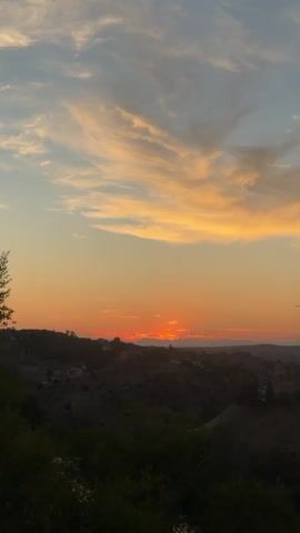 The video displays a panoramic view of a town situated on a hillside, bathed in the warm glow of a sunset. The sky is a gradient of colors, transitioning from a deep orange near the horizon to a soft blue overhead, with wispy clouds tinged in shades of pink and gold. The sun itself is partially obscured by the distant hills, casting a warm, diffused light over the scene. The town's buildings, a mix of traditional architecture, are clustered together on the slopes, with a prominent church spire visible in the middle distance. The foreground is dominated by dark, silhouetted vegetation, adding depth and contrast to the image. The overall impression is one of tranquility and natural beauty, capturing a moment of serene twilight over a European landscape.