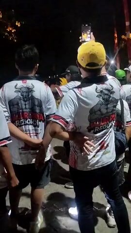 This video shows a nighttime outdoor scene with a crowd of people. In the foreground, two individuals are visible from the back, wearing matching white t-shirts with a graphic of a muscular bull and the text "BALIRAYA REBORN." One person is wearing a yellow cap and holding up a smartphone, possibly recording or taking a photo. Another person to their left is wearing a black cap and is also looking towards something ahead. In the background, other people are visible, some with their backs to the camera, and there are lights and what appears to be some kind of decoration or float. The overall atmosphere suggests a lively gathering or event.
