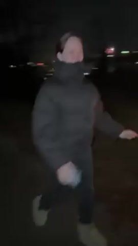 At night, a person wearing a black puffer jacket, dark pants, and light-colored boots runs away from a burning object in a grassy field. The person is smiling. In the background, city lights and fireworks are visible. The burning object emits sparks and smoke. The ground is covered in dark leaves and grass. Trees are sparsely visible in the background. The camera shakes as the person runs away.