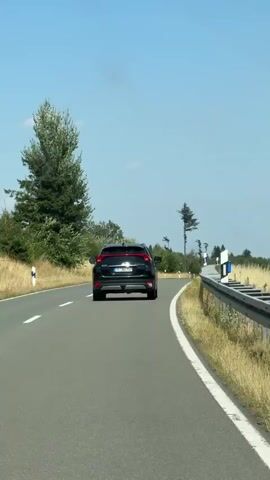 A black SUV is driving on a paved road with a white dashed line on the left and a solid white line on the right. There is a guardrail on the right side of the road, beyond which is dry grass and vegetation. On the left side of the road, there are more trees and vegetation. The sky is clear and blue. The camera is behind the SUV and appears to be moving at the same speed.