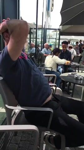 The video shows an outdoor cafe scene. In the foreground, a middle-aged man sits on a metal chair with his hands behind his head, leaning back. He wears a navy blue t-shirt, black pants, and has a visible belly. 

In the background, other people are seated at tables. There are two older men seated together at a table, and a younger man and girl seated at another table. There are cigarettes and ashtrays on the tables. 

The cafe is beside a building with large glass windows that reflect the surrounding area. Some umbrellas are visible, providing shade. The ground is paved. The overall atmosphere is casual and relaxed.