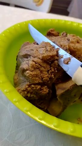 A close-up shot shows a person's hand holding a knife and cutting a piece of meat in a green bowl. The meat appears to be cooked and is brown in color. The knife has a blue and white handle. The bowl is placed on a white surface. In the background, a partially visible object with a pattern of yellow bananas can be seen. The lighting is natural, and the focus is on the meat and the knife.