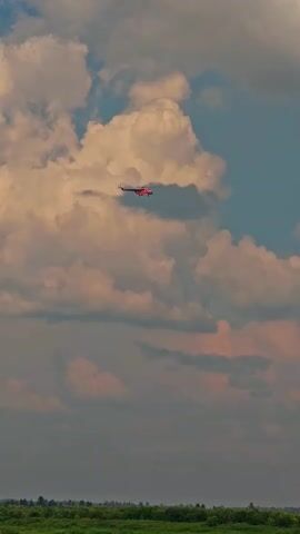 A red helicopter flies across a pale blue sky with fluffy white and pink clouds. Below the clouds, a dark green, forested landscape is visible. The helicopter is a small, distant object in the frame.