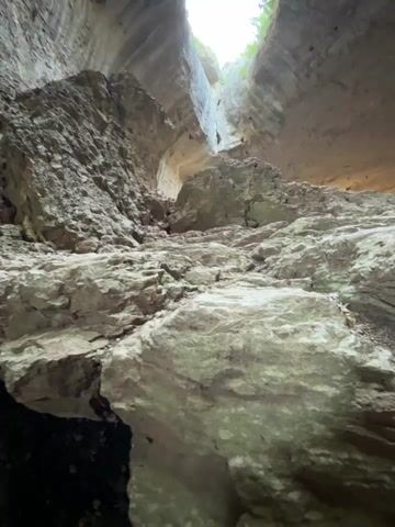 The video is a low-angle shot looking up into a massive cave or sinkhole. The cave walls are rough and textured, appearing to be made of rock. There are at least two large openings in the ceiling of the cave, through which bright daylight is visible. Green foliage can be seen around the edges of these openings, indicating that the cave is in an outdoor environment. The foreground of the shot is dominated by large, irregular rock formations, suggesting a collapsed or eroded area within the cave. The perspective gives a sense of immense scale and natural grandeur. There are no people or distinct objects visible, only the natural geological formations of the cave and the light from above.