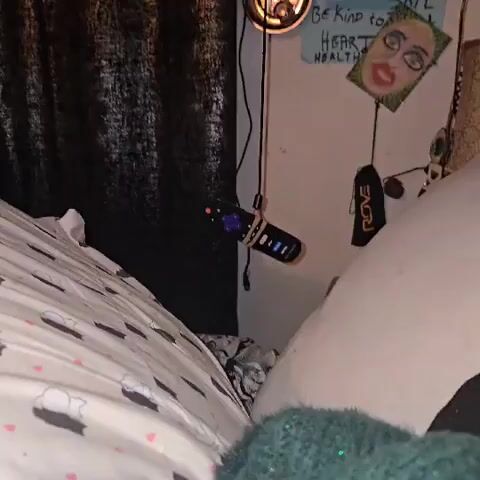 The video shows a close-up of a Roku TV remote hanging from a cable next to a bed. The remote is black with a purple directional pad and buttons for Netflix, Apple TV+, Paramount+, and HBO Max. It is secured to the cable with two rubber bands. The bed has white sheets with a pattern of gray clouds and pink hearts. A green, fuzzy sock is visible at the bottom of the frame. To the left, there is a black curtain. On the wall, there is a small light fixture and a few pieces of decor, including a cutout of a face and a sign that reads "Be kind to your HEART HEALTH."