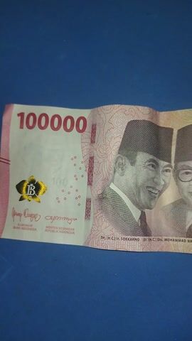 The video shows a close-up, top-down view of an Indonesian 100,000 rupiah banknote placed vertically on a blue surface. The banknote is folded horizontally, with the top portion showing the faces of two men. The man in the foreground is wearing a traditional cap and smiling, while the man behind him is wearing glasses and also smiling. The denomination "100000" is clearly visible in red lettering on the lower part of the banknote, along with a gold holographic emblem and text that appears to be in Indonesian. The banknote is slightly wrinkled. The background is a solid, dark blue color. There are no people or other objects visible in the scene.