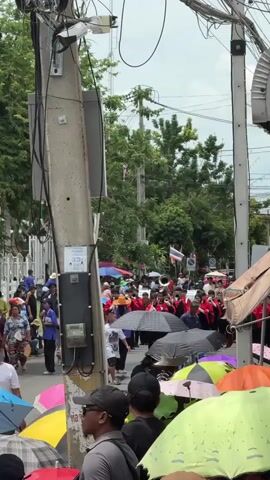 The video shows a street scene with a parade or procession. In the foreground, several people are visible holding colorful umbrellas, suggesting it may be sunny or the threat of rain. One person in the foreground is wearing a gray t-shirt and a black baseball cap. To the left, a utility pole with numerous wires and some electrical equipment is prominently displayed. Further down the street, a group of people in matching red and black uniforms are marching. Some are playing instruments, including what appears to be a brass band. They are preceded by individuals in blue shirts and hats. The street is lined with trees and buildings, and other spectators are gathered along the sides of the road, some holding umbrellas as well. The overall atmosphere appears to be a public event or celebration.