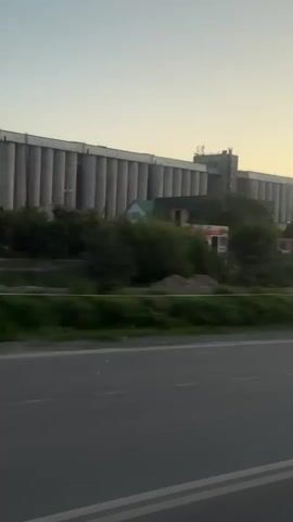 This video captures a view from a vehicle moving along a paved road with double white lines. The scene is dominated by a long, multi-story industrial building with a row of cylindrical silos on its left. Several smaller buildings and trees are visible in front of the industrial complex. The sky is clear with a pale orange hue, suggesting it might be dawn or dusk. The perspective suggests the camera is looking slightly upwards and to the right, with part of a vehicle's side mirror visible in the bottom right corner. The overall environment appears to be an industrial or semi-urban area.