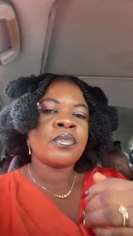 A close-up shot shows a Black woman in a car, wearing an orange top and makeup. She has her hair styled in large twists. She touches her face and adjusts her earrings while looking into the camera. The background is blurred, but shows the interior of a vehicle.