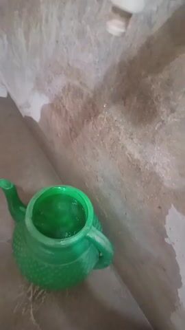 A close-up, high-angle shot shows a green, plastic, teapot-shaped bucket placed on a tiled floor. The bucket is partially filled with water, which appears slightly green, possibly reflecting the bucket's color. Directly above the bucket, a water tap is visible, from which water is slowly dripping. The background consists of a mottled, light brown and grey tiled wall, showing signs of wear or staining. The setting appears to be a utilitarian space like a bathroom or washing area in Kakki, Pakistan. No people or text are visible.