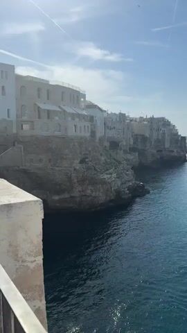 The video captures a panoramic view of an idyllic coastal scene, opening with a shot of ancient stone buildings perched on cliffs overlooking a vibrant blue sea. As the camera pans right, it reveals the vast expanse of the ocean under a clear sky dotted with sparse clouds. Several boats are seen traversing the tranquil waters, leaving gentle wakes in their paths, while a single yellow buoy bobs placidly in the distance. The sun glints off the water, creating a sparkling effect, and the overall atmosphere is one of serene beauty.