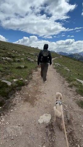 A person walks up a dirt path on a grassy hillside. The person is wearing a dark hooded jacket, brown pants, and a backpack. A small, light-colored dog on a leash walks beside them. The path curves to the right and leads toward distant mountains. The sky is blue with scattered white clouds.