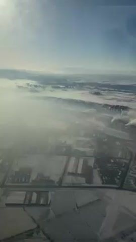 The video presents an aerial view of a landscape covered in snow and fog. The scene is dominated by a layer of fog obscuring much of the land below, with a faint blue sky visible above. In the foreground, there are patches of snow-covered fields or plots of land, arranged in a grid-like pattern. Structures resembling buildings or industrial facilities, also covered in snow, are scattered throughout this area. In the distance, mountain ranges can be seen emerging from the fog, with what appears to be an industrial area or power plant emitting plumes of smoke. Overall, the scene evokes a sense of coldness, desolation, and possibly environmental pollution.