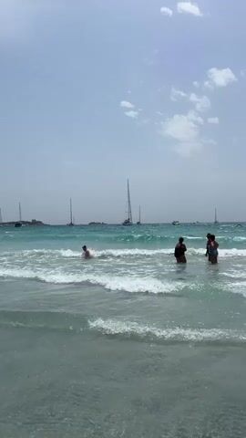 The video shows a beach scene with people in the ocean and sailboats in the distance. The water is clear and blue, with waves breaking on the shore. There are several people wading in the water, some of whom appear to be children. In the background, several sailboats are visible on the horizon. The sky is clear and blue with a few scattered clouds. The overall impression is one of a peaceful and relaxing day at the beach.