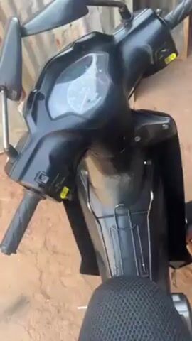 The video shows the front of a black scooter. The scooter has a speedometer with visible kilometer per hour markings. The handlebars and mirrors are visible. The scooter is parked on a reddish-brown, potentially earthen, surface. A corrugated metal wall is partially visible in the background. The overall scene is shot from a slightly high angle, focusing on the front section of the scooter.