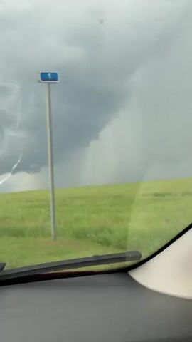 The video shows a view from inside a vehicle, looking out through the windshield at a landscape. The sky is overcast with dark, ominous clouds, and a visible shaft of rain is falling in the distance over a green field. The windshield wipers are visible in the foreground. The overall impression is that of an approaching storm.
