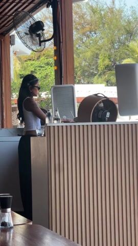 A woman wearing sunglasses, a white tank top, and black pants stands behind a countertop, facing a window in a tropical-looking restaurant. She has long, dark hair and a tattoo on her left arm. On the countertop are a water bottle, a glass, a square air purifier, a round fan, and a computer monitor. A large ceiling fan spins above. Outside the window are trees and possibly buildings. The countertop has a light-colored vertical slat pattern. A table with glasses and a pepper shaker are in the foreground.