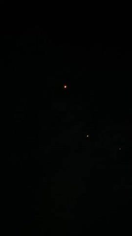 The video shows a dark sky with a few bright, reddish-orange lights. One light is larger and brighter than the others and is located towards the top center of the frame. Two smaller lights are visible below and to the right of the largest light. The lights appear to be stationary against the dark background, with faint cloud-like formations visible in the darkness. The video is brief and without sound.