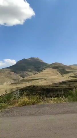 The video is a landscape shot looking towards a mountain range. The foreground consists of a dirt road and a yield sign. The middle ground features rolling hills with terraced fields and sparse vegetation. The background is dominated by a large mountain under a clear blue sky with scattered clouds. The camera appears to be moving slightly, panning from left to right. The overall impression is one of a rural, mountainous environment.