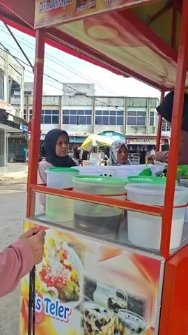 The video shows a street food cart with a red metal frame and a glass display case. Inside the display case are several white and green plastic containers, some filled with liquids. A sign on the front of the cart depicts an image of a colorful dessert labeled "Es Teler" and another image of layered drinks. Two women are standing behind the cart, and one person's arm is visible on the left side of the frame, reaching towards the cart. The background shows a street with buildings and other stalls, suggesting an outdoor market or commercial area. The lighting indicates it is daytime.