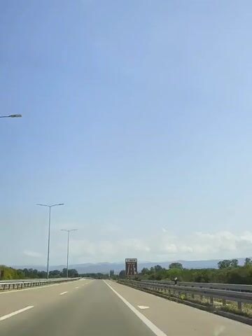 This is a first-person perspective video taken from a vehicle traveling on a highway under a clear blue sky. The road has multiple lanes, marked with white lines, and a guardrail lines the right side of the road. On the left, there are streetlights. In the distance, there are trees and hills, with a billboard on the right side of the road. There are no other vehicles or people visible.