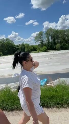 A young man records a selfie video while walking with a woman, presumably his partner, along a paved path. The man wears a white t-shirt with a Nike logo, a baseball cap, and has a beard. He looks at the camera and smiles. The woman wears a white and grey striped shirt, shorts, sunglasses, and hoop earrings. She points to the right, toward a large field covered with white plastic or material. The sky is blue with fluffy white clouds, and green trees are visible in the background. The man points to the right as well. The pair are walking along the path beside a patch of green grass.