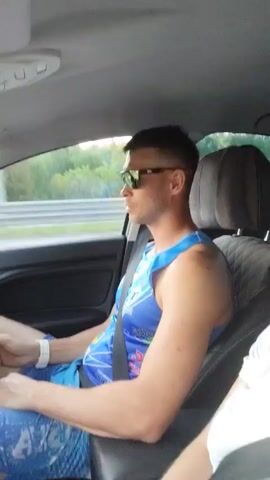 The video shows a man and a woman driving in a car. The man is driving, wearing a blue athletic tank top and sunglasses. The woman is in the passenger seat, wearing a white t-shirt. She is talking into the camera, and the car appears to be traveling down a highway with a forest visible in the background. Music plays in the background.