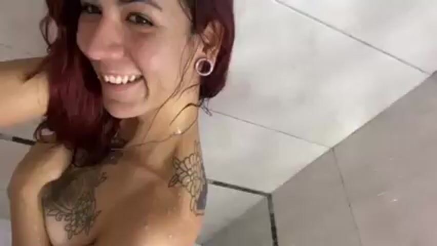The video shows a smiling woman with red hair in a shower. She has tattoos on her chest and shoulder, and ear plugs in her ears. Water is running over her face and body. The shower has grey and white tiled walls.