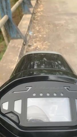 The video begins with a close-up of a scooter's digital dashboard, showing a speed of 0 km/h and a time of 5:58. The camera then pans to the right, revealing a concrete bridge with blue railings and a tree-lined landscape. As the video progresses, the camera moves forward, showing a stream flowing through a rocky bed surrounded by lush vegetation, including palm trees and banana plants. The scene appears to be in a rural or tropical area. There are no people visible in the video.
