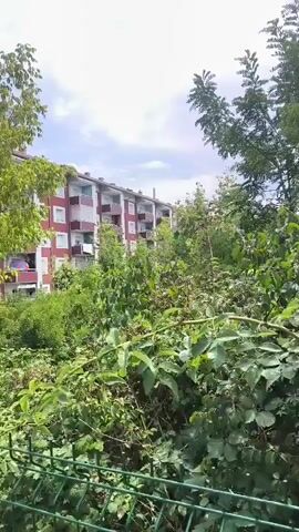 The video shows a view of apartment buildings from behind a wire fence and dense foliage. The buildings are painted in shades of red and white. The camera then pans to show a train track next to a paved walkway. A tall, pink pole with a sign is visible on the right side of the walkway. The camera continues to pan, showing more of the train tracks and apartment buildings in the background, surrounded by trees and overgrown vegetation. The sky is blue with some clouds.