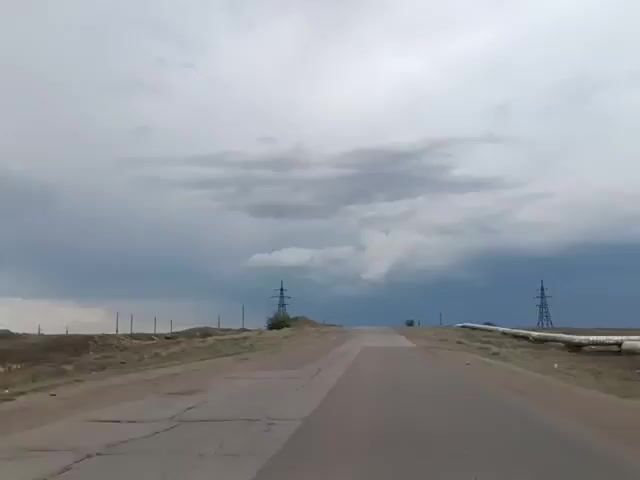 A short video shows a car driving on a paved road. The road is cracked and appears to be in disrepair. The landscape on either side of the road is barren and dry, with sparse vegetation. There are power lines and pylons visible in the distance. The sky is overcast with dark, dramatic clouds, suggesting an impending storm. The video captures the feeling of a journey through a desolate and atmospheric landscape.