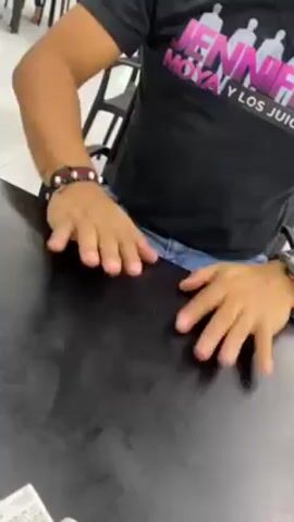 A person is seated at a black table, wearing a black t-shirt with a graphic of people and the words "JENNIFER MOYA Y LOS JUICES." The person is also wearing blue jeans and bracelets. Their hands are resting on the table and making a drumming motion. Other people can be seen out of focus in the background.