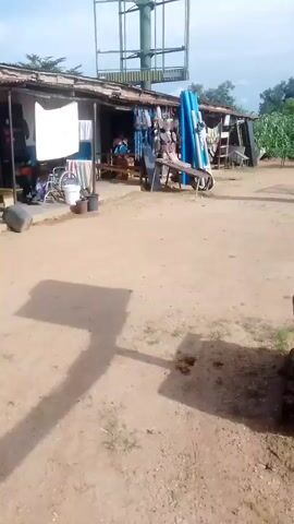 This media file captures a view of an outdoor marketplace stall in Unguwar Saya, Nigeria. The scene is dominated by a makeshift structure with a corrugated iron roof, likely serving as a storefront. Various items are displayed for sale, including what appear to be clothing, footwear, and potentially household goods. Among the objects are a wheelchair, buckets, and bundles of blue pipes leaning against the stall. A tall, metal billboard-like structure stands prominently in the background, its purpose unclear in this context.

The ground is sandy, typical of a dry, outdoor environment. The lighting suggests it is daytime, with shadows indicating the sun is at a moderate height in the sky, possibly late morning or early afternoon. The sky is partly cloudy. The overall impression is of a working marketplace, likely serving a local community. There are no explicit indications of a specific event or festival taking place, but rather a snapshot of daily commerce. The presence of the wheelchair might suggest that some patrons or owners have mobility challenges, or that such items are also sold. The environment appears somewhat rustic and informal. No legible text is visible in the media file.
