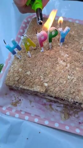 A hand holding a green lighter is igniting candles on a rectangular cake. The candles are shaped like letters that spell out "HAPPY". The cake is covered in crumbled pastry and appears to have layers with a pink filling. It is placed on a pink and white polka-dot tray. The background is blurred but suggests an outdoor or casually decorated setting. The action is focused on the process of lighting the birthday candles.