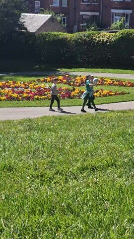 Two children and one adult are walking on a sidewalk in a grassy park. The sidewalk has a flower bed along one side. The adult is wearing a light colored top and dark pants. One child is wearing a green top and dark pants. The other child is wearing a dark top and pants. The children are throwing a ball. Brick buildings are visible behind a hedge along one side of the park.